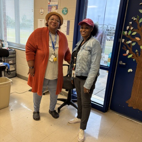 Lady in red dress and lady and jean jacket, both wearing hat hats in front of the window