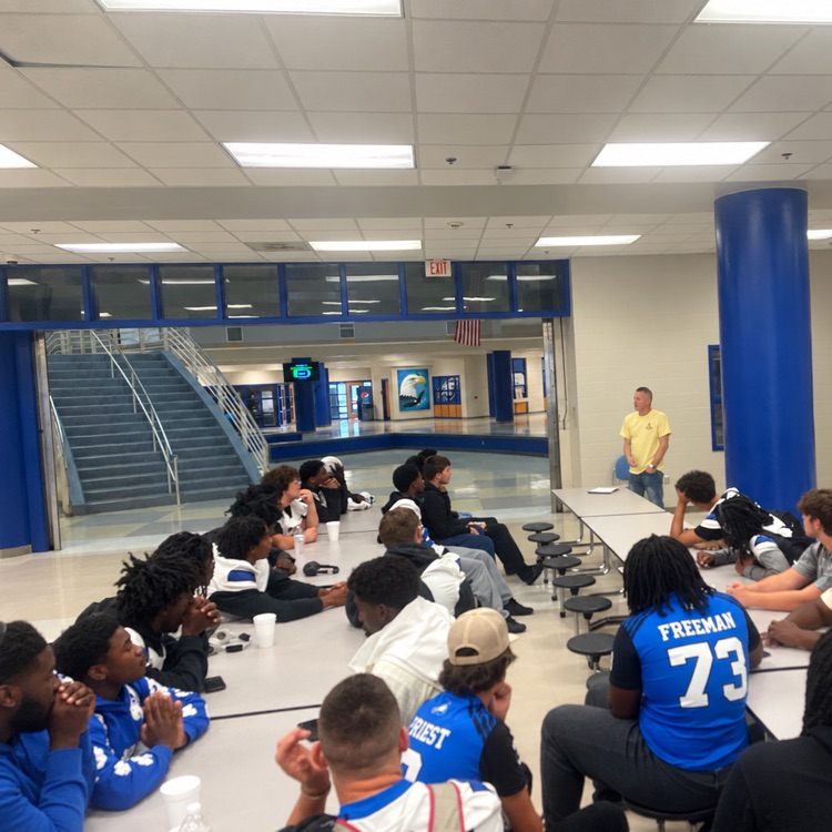 large group of football players at cafeteria tables listening to someone talk to them before they eat
