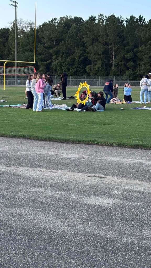 Students gather on blankets across a grassy football field next to a sun-shaped Senior Sunrise cutout.