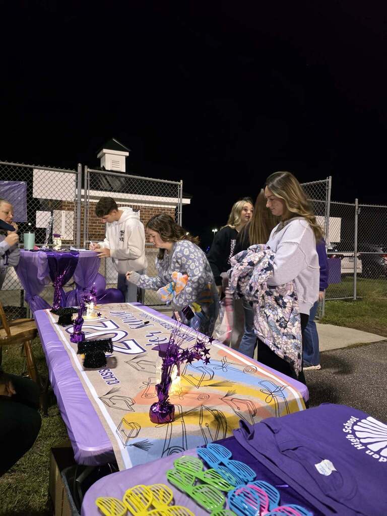 Students gather outdoors at night around tables decorated with purple banners and graduation crafts for Senior Sunrise. 