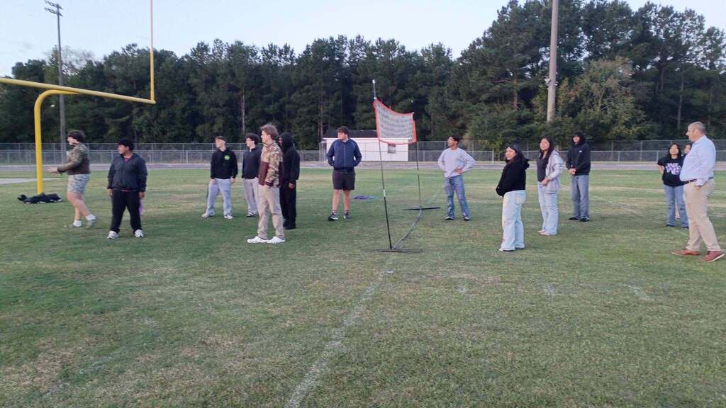 Students stand on a grassy football field near the end zone and goalposts during an outdoor morning event.
