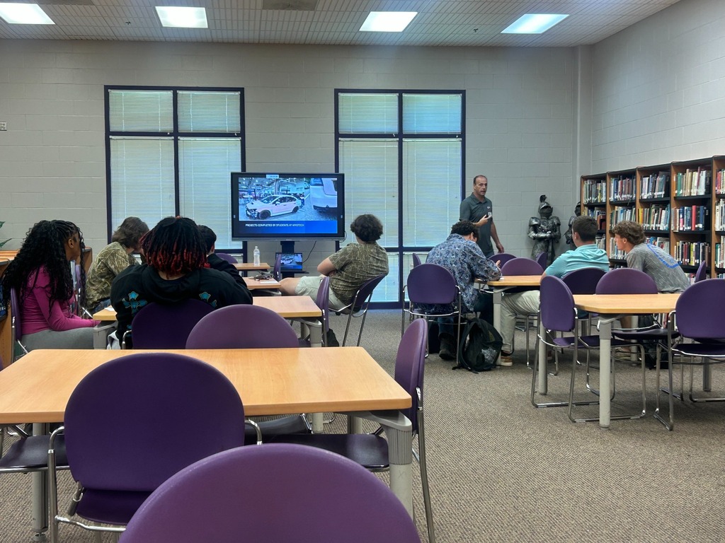 A WyoTech representative speaks to students seated at tables in the school media center while a presentation is displayed on a screen.