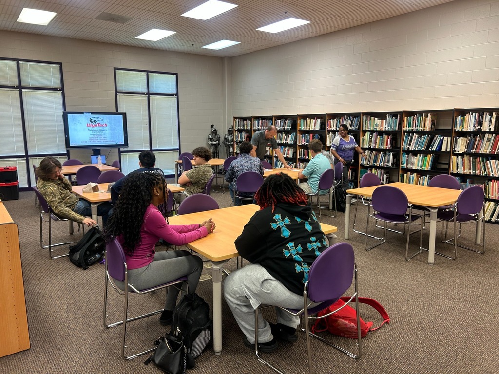 Students sit at tables in the school media center before or during a WyoTech information session, with a WyoTech presentation displayed on a screen at the front of the room.