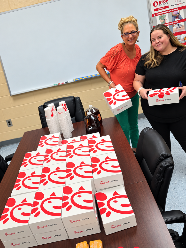 1: Two BMS staff members smiling while holding Chick-fil-A catering boxes beside a table filled with boxed lunches, drinks, and cups.
