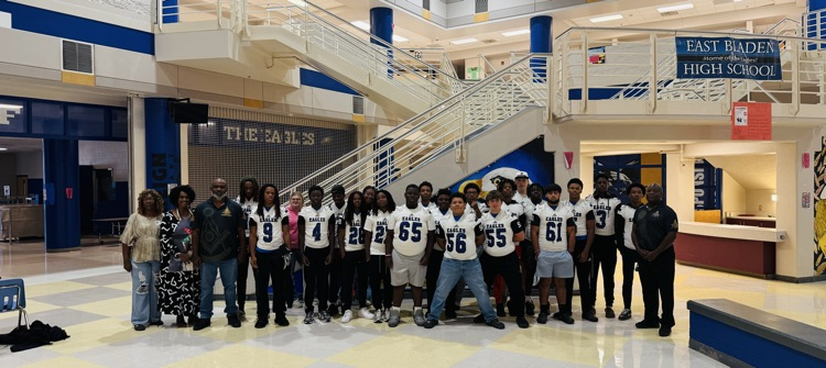 Group of football players and two gentlemen and two women stand together in front of stairs in the atrium of East Bladen