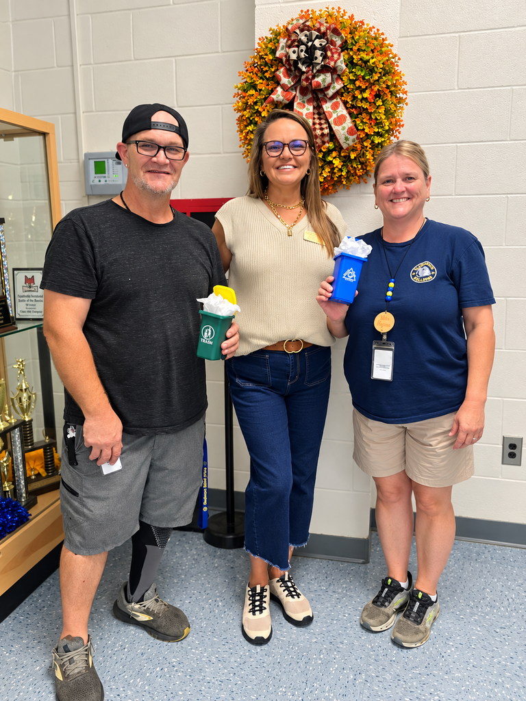 Alt text: Three smiling BMS staff members pose together in front of a fall wreath while celebrating Custodian Appreciation Day. Mr. Pait and Mrs. Ransom hold small trash and recycling containers as appreciation gifts.
