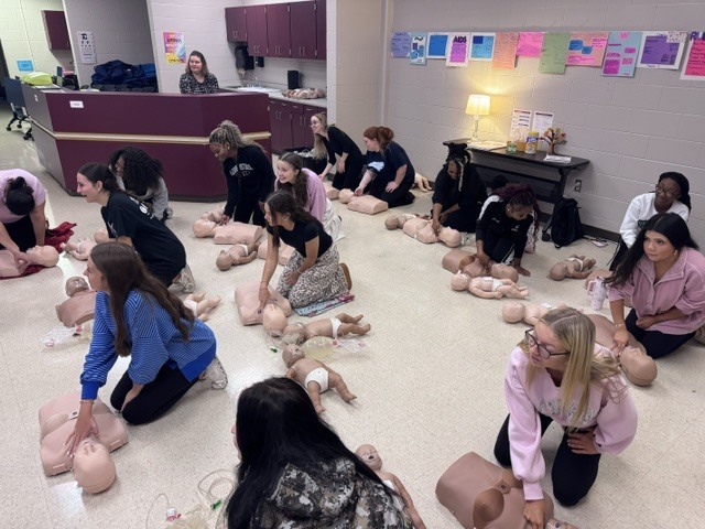 Students practicing infant CPR on training mannequins on a classroom floor during a health class.