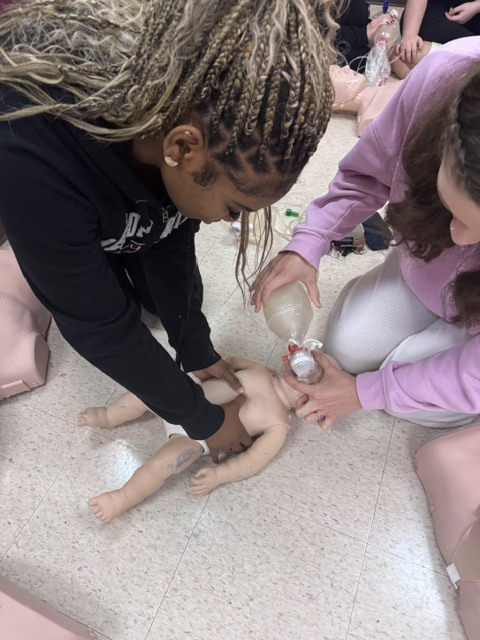 Two students demonstrate proper CPR technique and bag valve mask ventilation on an infant mannequin.