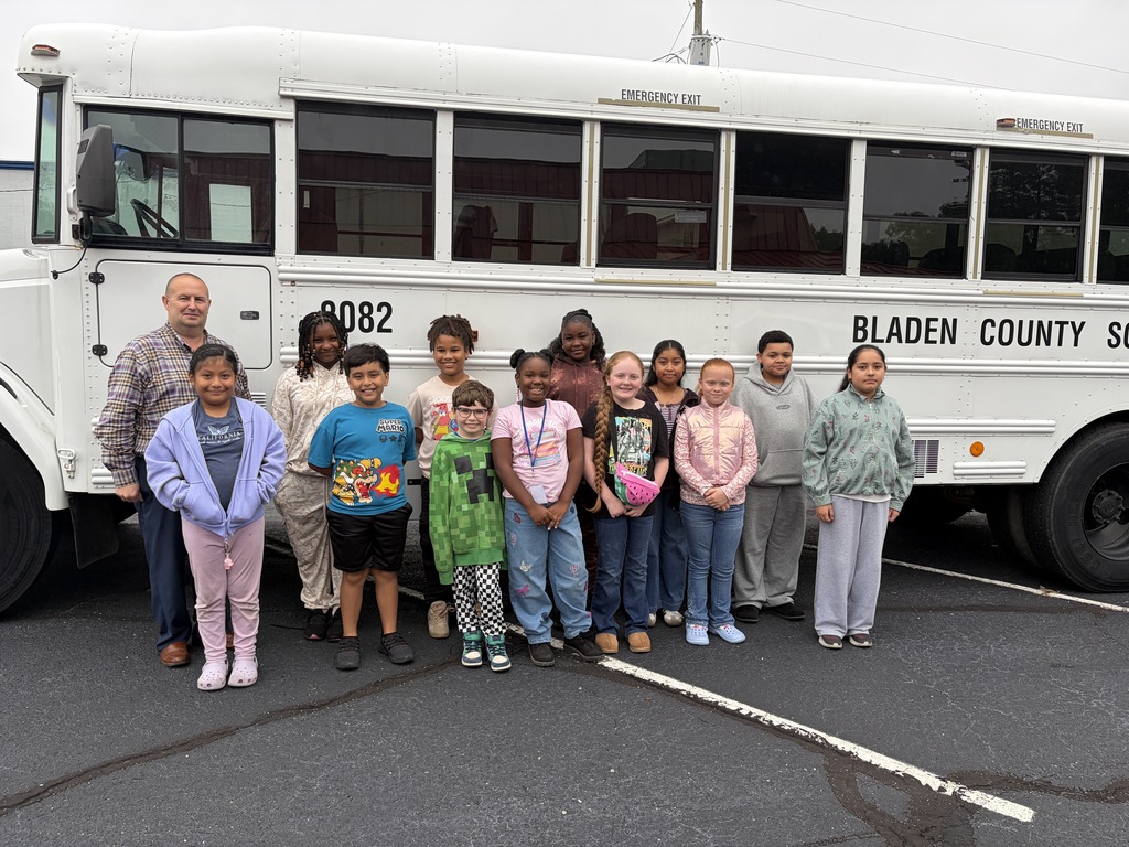 Elementary student advisory students stand smiling with the superintendent in front of a Bladen County Schools activity bus.