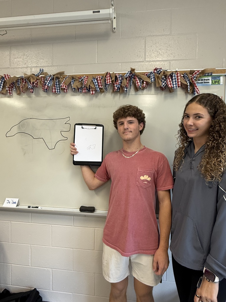 Two students standing in a classroom; one holds up a clipboard with a drawing, next to a whiteboard outline of North Carolina.