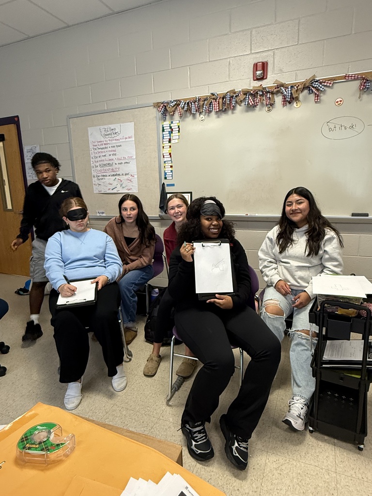 Students in a classroom, including two wearing blindfolds, holding clipboards during a group activity.