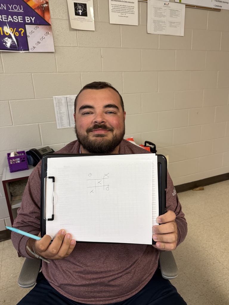 A smiling teacher holding up a clipboard with a piece of paper inside a classroom.