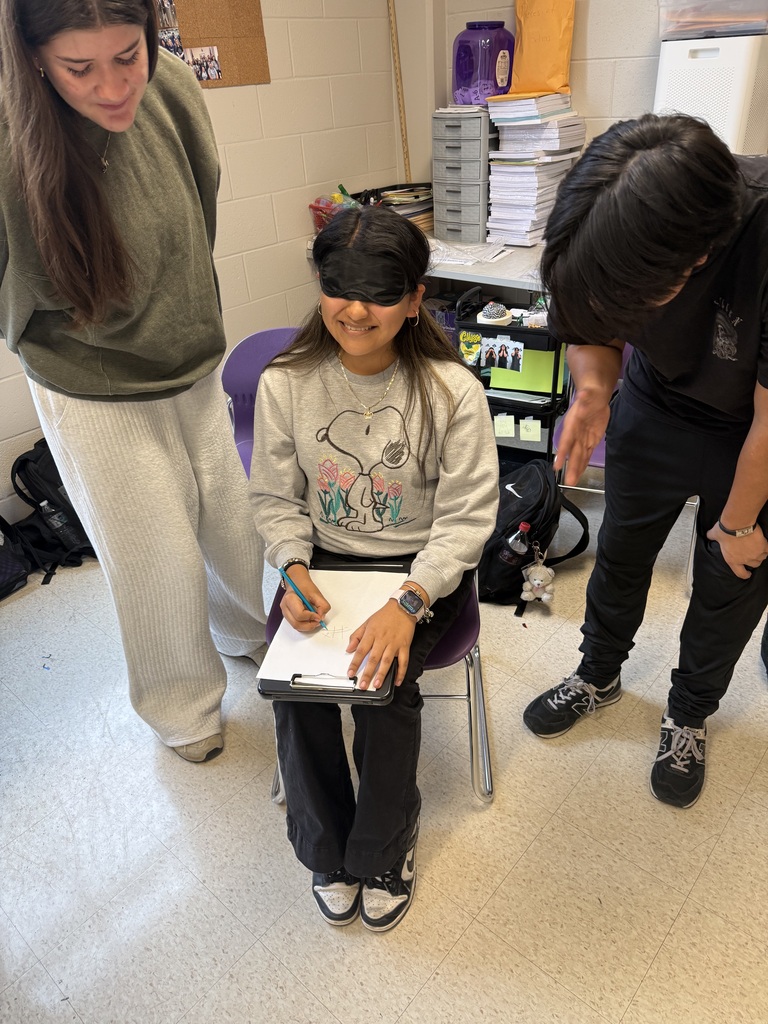A smiling blindfolded student wearing a Snoopy sweatshirt sits as two classmates guide her during a classroom activity.