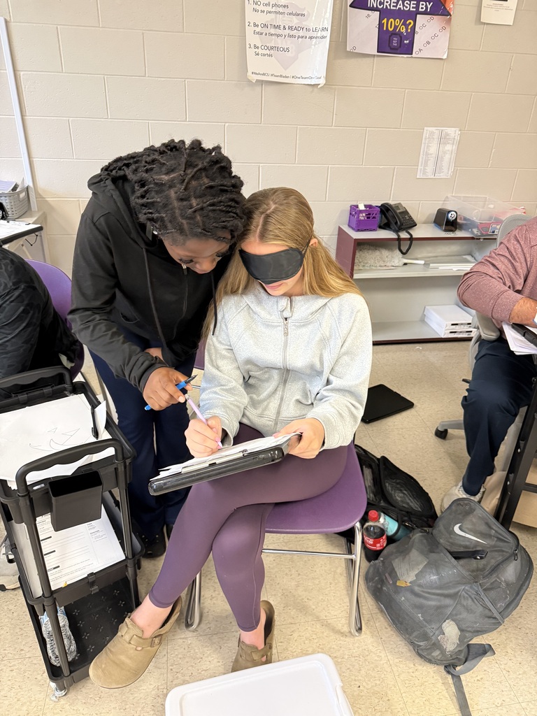 A classmate leaning over to assist a blindfolded student as she draws during a classroom activity.
