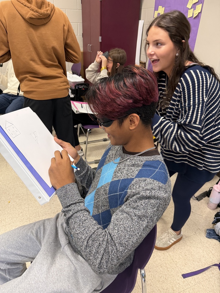 A student with red-highlighted hair wearing a blindfold draws a house while a smiling classmate coaches him