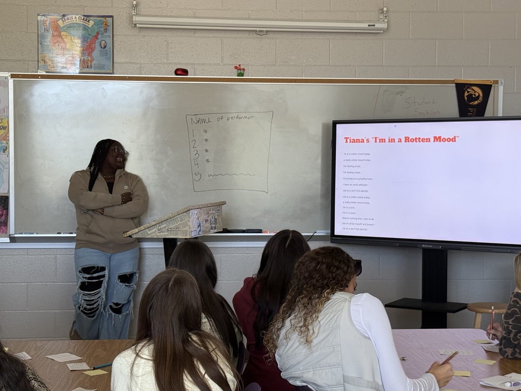 A smiling presenter standing near a whiteboard and digital display as students watch from their desks