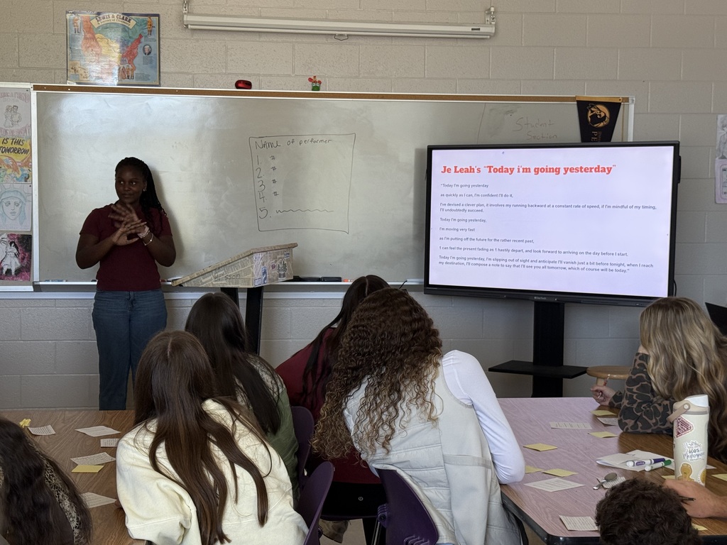 A presenter stands by a whiteboard and display screen, speaking to seated students in a classroom.
