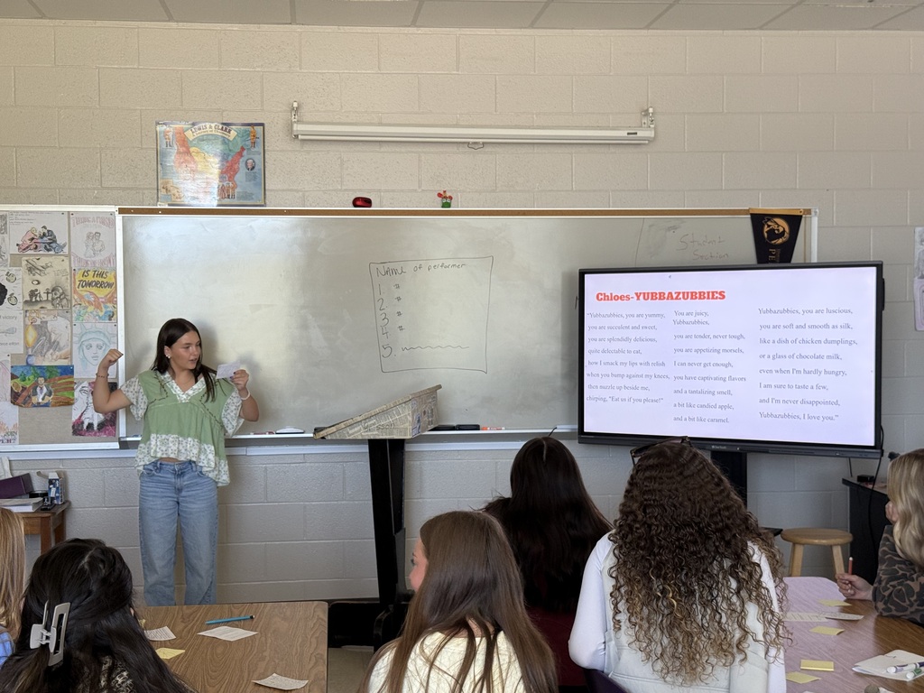 A presenter gestures with a paper in hand near a digital display screen as students look on in class