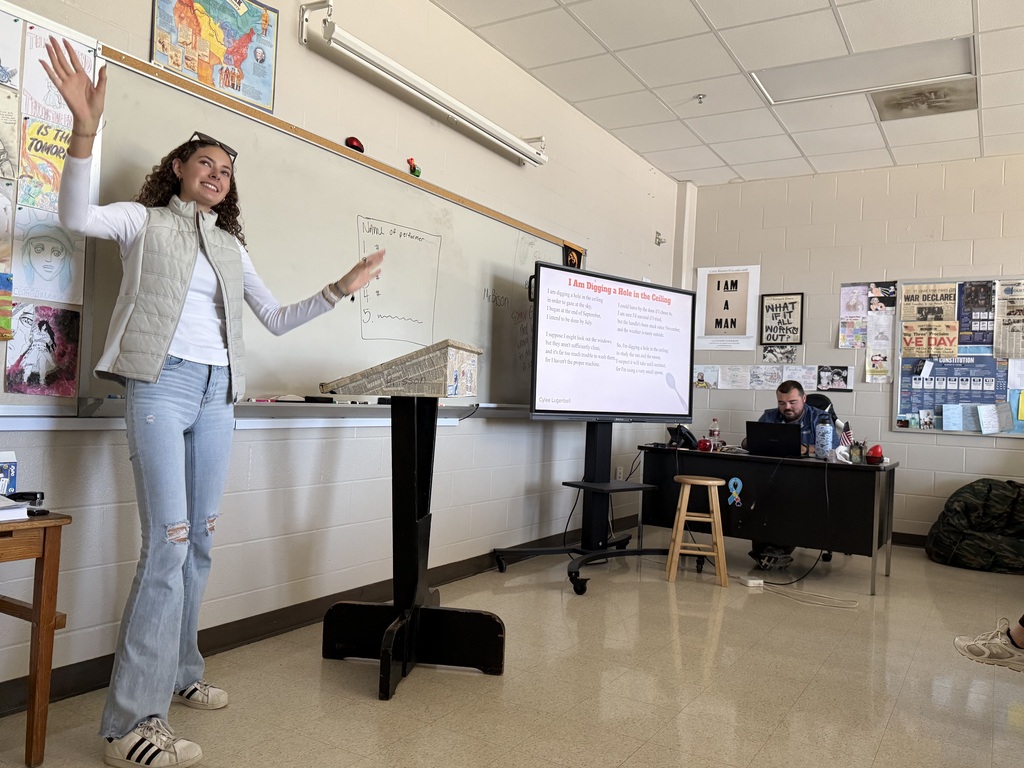 A presenter gestures with arms raised near a whiteboard and a display titled "I Am Digging a Hole in the Ceiling"
