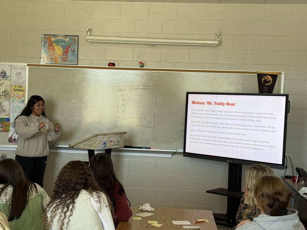 A presenter gestures with both hands near a whiteboard and a display showing text titled "Melissa 'Oh, Teddy Bear'"