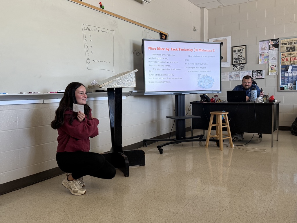 A student kneels holding a card near a podium while a screen displays text titled "Nine Mice by Jack Prelutsky