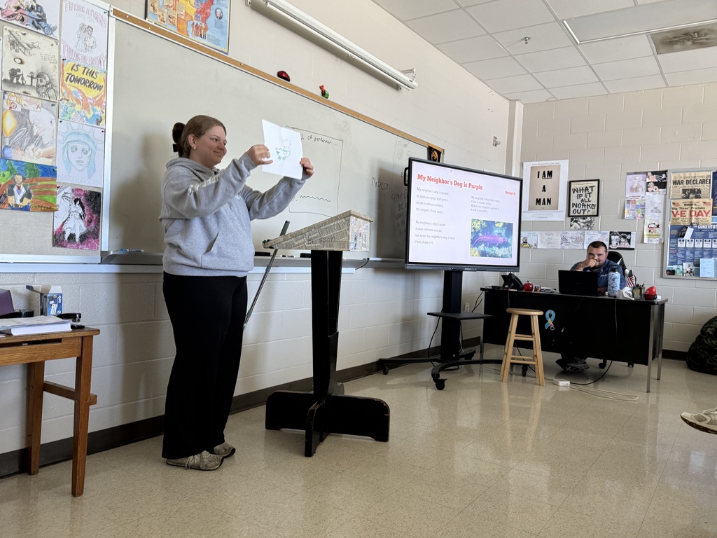 A presenter holds up a sheet of paper with a drawing near a podium and a display titled "My Neighbor's Dog is Purple