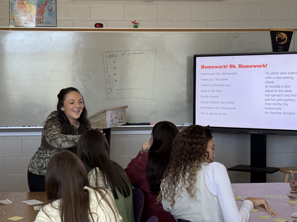 A student smiling and handing an object to students seated at a table in front of a classroom whiteboard