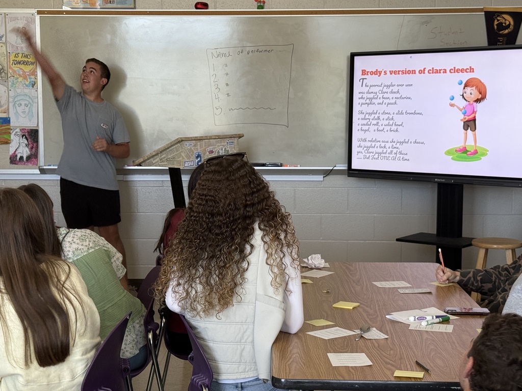 A male student gestures toward a poster near a whiteboard while a display shows text titled "Brody's version"