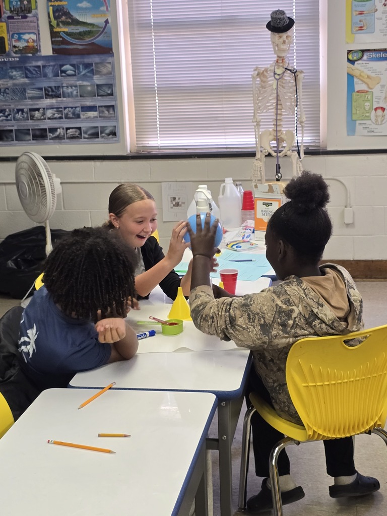 Three Bladenboro Middle School fifth graders excitedly observe a hands-on chemical reaction experiment involving a blue balloon. Students collaborate around a classroom table with science materials, demonstrating curiosity, teamwork, and learning through discovery.