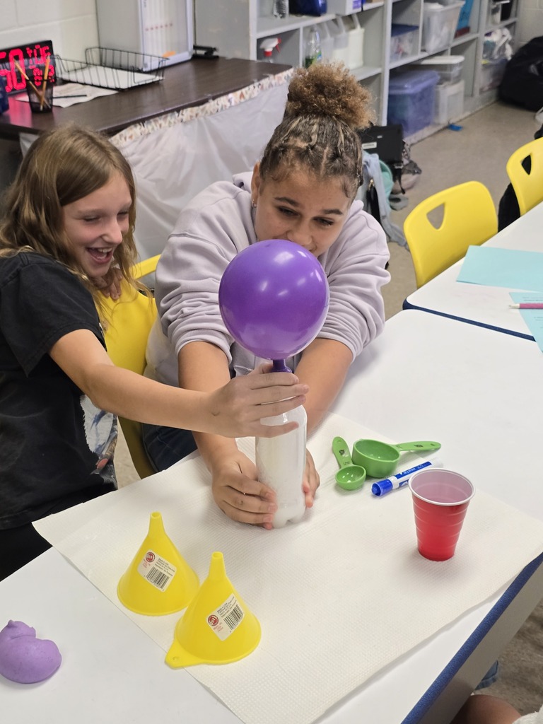 Two Bladenboro Middle School fifth graders excitedly observe a purple balloon inflating during a hands-on chemical reaction lab. Students explore how mixing substances produces gas, demonstrating changes in matter through scientific observation and discovery.