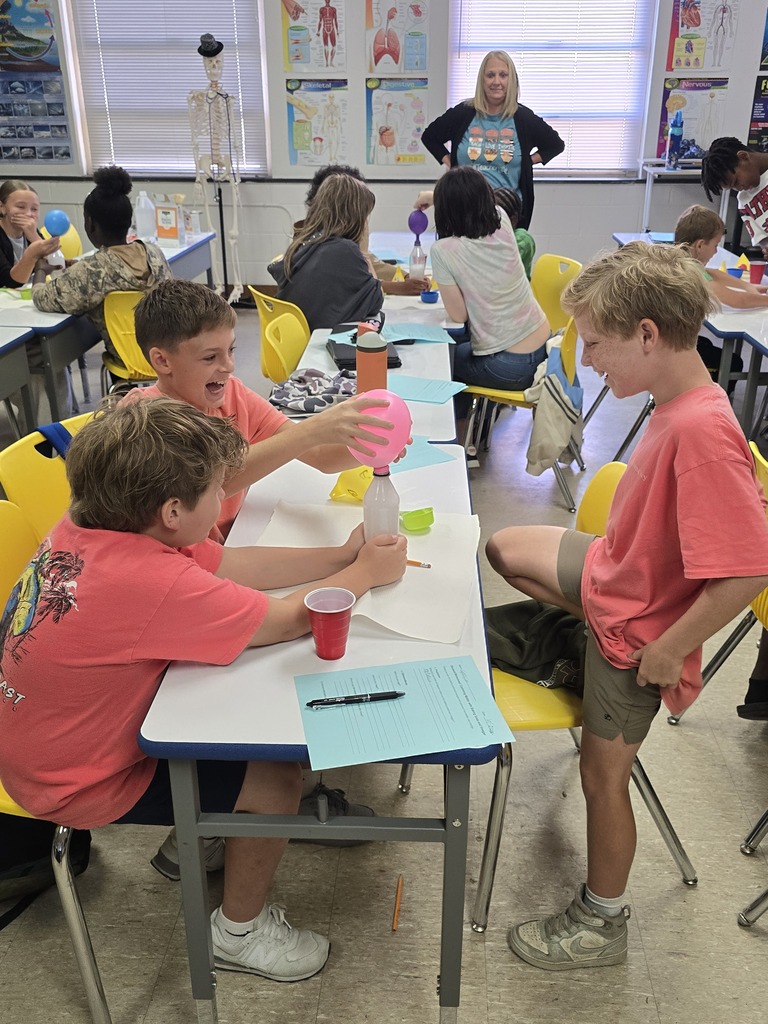 Bladenboro Middle School fifth graders enthusiastically participate in a hands-on chemical reaction lab. Three students in the foreground excitedly observe a pink balloon inflating on a bottle as classmates conduct similar experiments. Their teacher supervises as students explore gas production, changes in matter, and scientific observation.