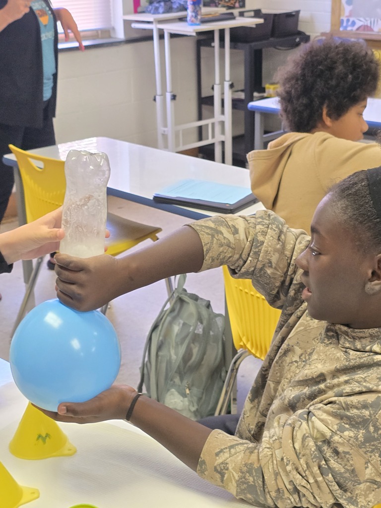 Bladenboro Middle School fifth grader excitedly displays a fully inflated blue balloon attached to a bottle during a hands-on chemical reaction lab. The student observes the foamy reaction inside the bottle, demonstrating how chemical reactions can produce gas and cause observable changes in matter.