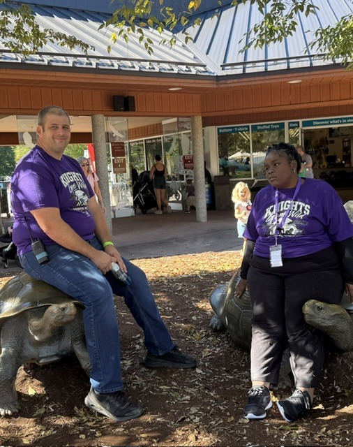 Female student and principal in purple tshirts at the zoo, sitting on turtles in the shade at the exit of the zoo