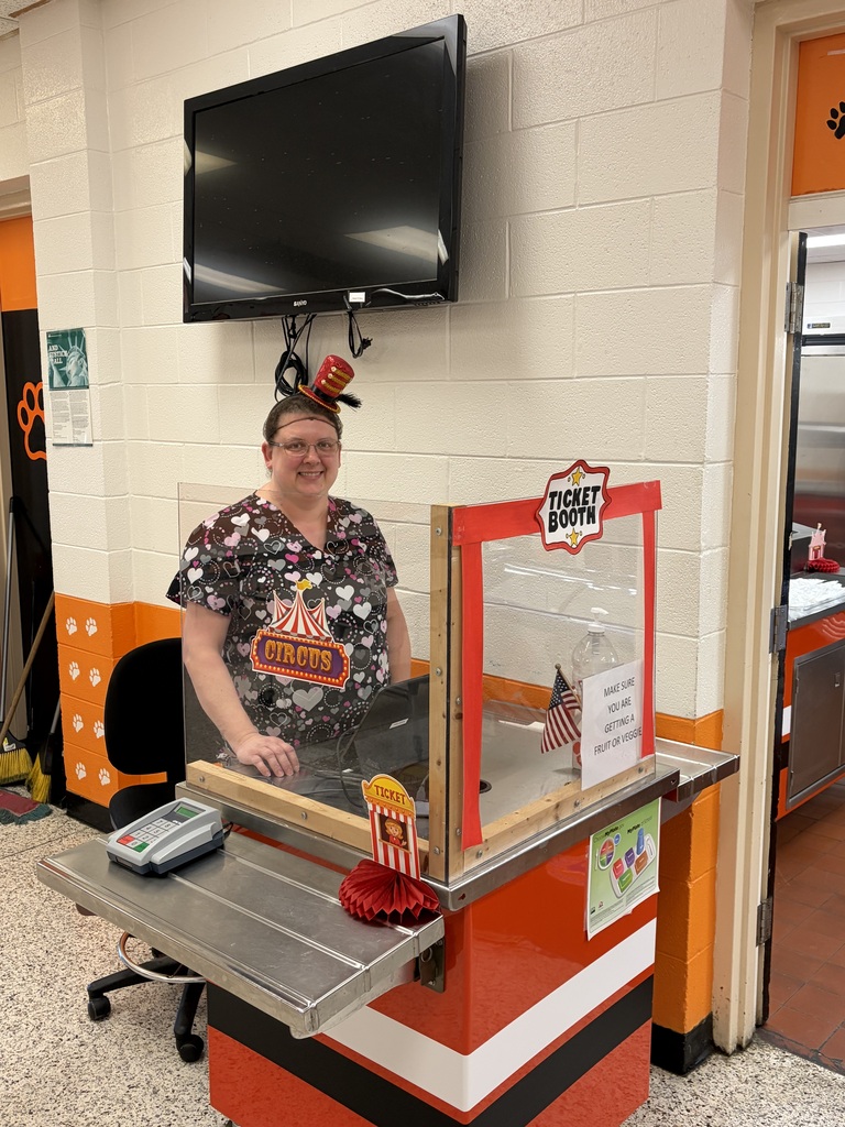 A cafeteria team member stands smiling behind the checkout counter that's decorated as a circus ticket booth.