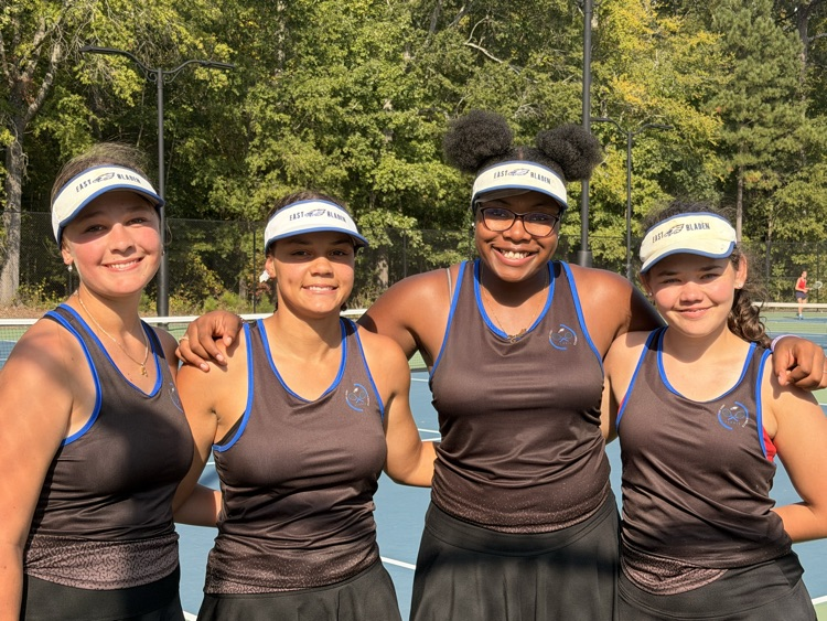 Group of four girls and black tennis skirts and tennis hats posing with one another on a tennis court