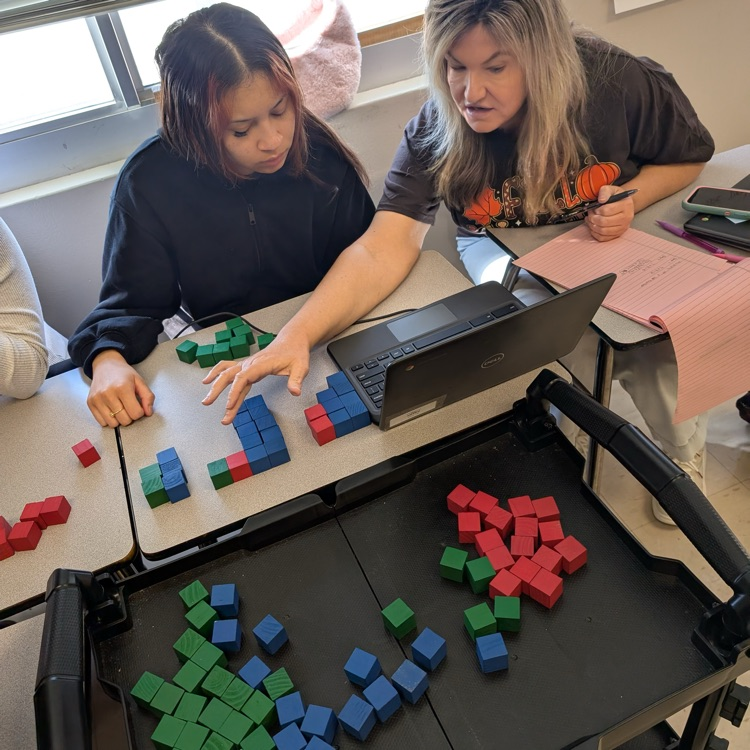 A cart with red blue and green blocks in front of a table with the same blocks as a teacher and student work