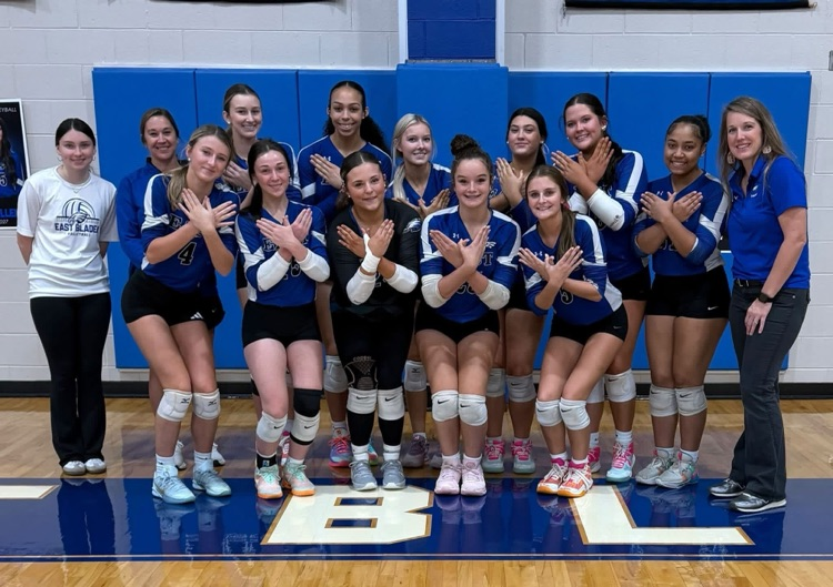 Group of volleyball players making a eagle wing sign with their hands in front of a padded wall on a basketball court