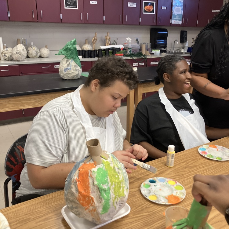 two students seen painting, papier-mâché pumpkins