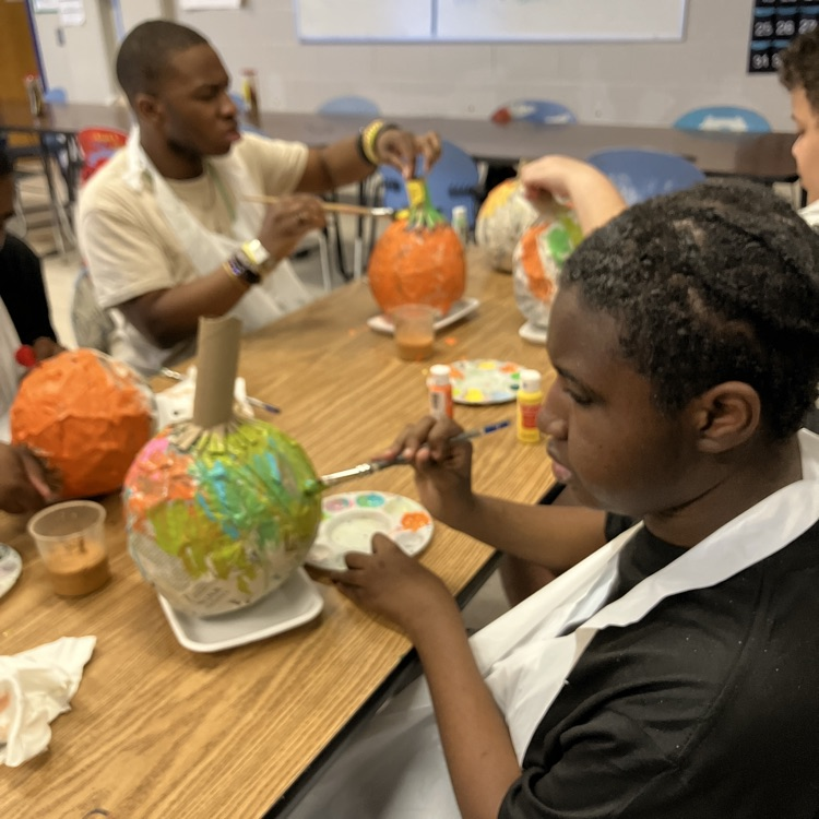 two students seen painting pumpkins that are made out of papier-mâché