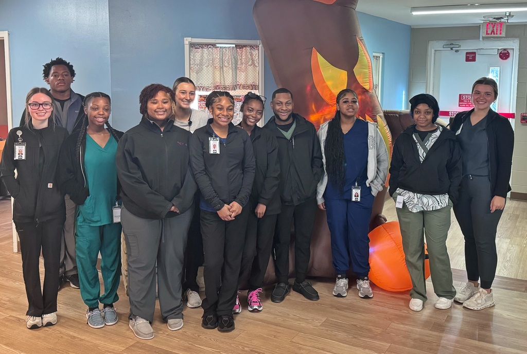 A group photograph of twelve diverse students in various colored medical scrub uniforms and lanyards, standing close together and smiling at the camera in an indoor setting. Behind them, to the right, a large inflatable holiday-themed decoration is partially visible.