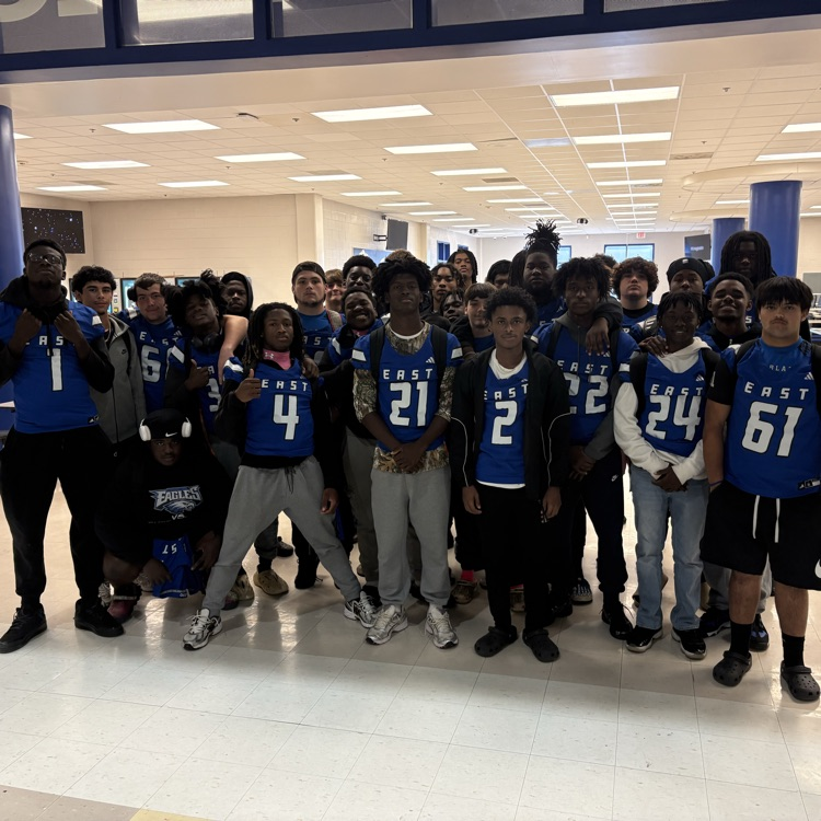 Group of football players standing in giving the thumbs up sign arms wrapped around one another person for a photo in front of the cafeteria