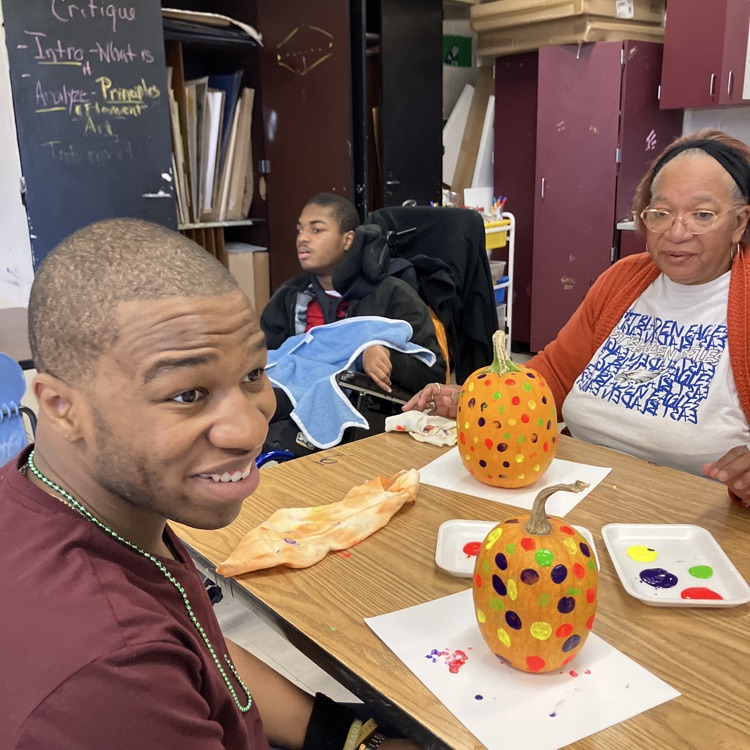 two students sitting and a teaching standing, all smiling, in an art room with paint a dotted pumpkins on the table