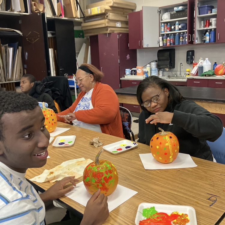 two students sitting and a teaching standing, all smiling, in an art room with paint a dotted pumpkins on the table