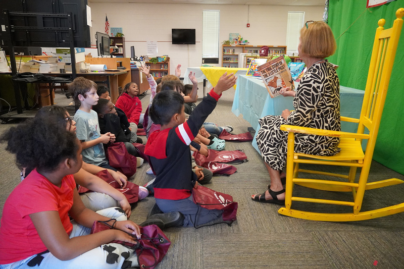 An adult sits in a yellow rocking chair reading books to a group of students.