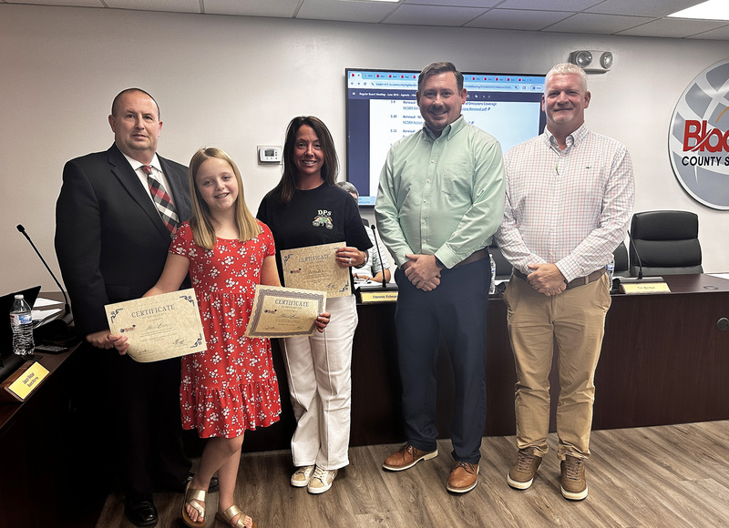 An elementary student and school staff stand with the Superintendent and Board Chair.