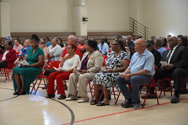 A group of adults sit in a gymnasium during a school event.