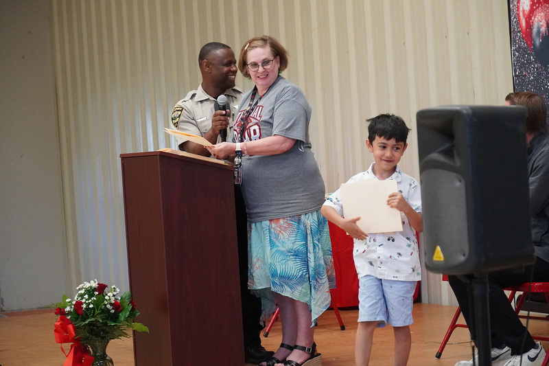 A teacher stands on stage at the podium and beside her is an elementary student who has just received a certificate.