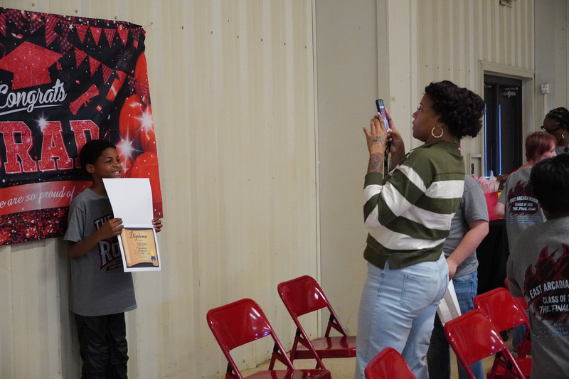 A parent is taking a photograph of a student in front of a Congrats Grad banner. The student is smiling and holding a certificate.