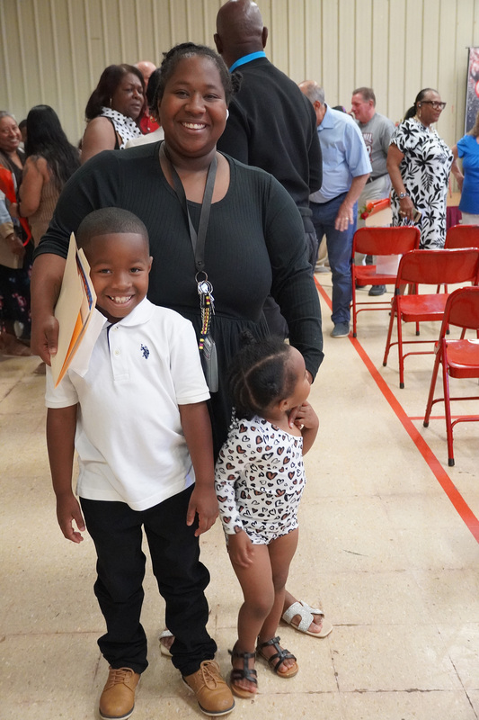 A parent stands smiling with her elementary student. A younger child is standing with them, but turned away from the camera.