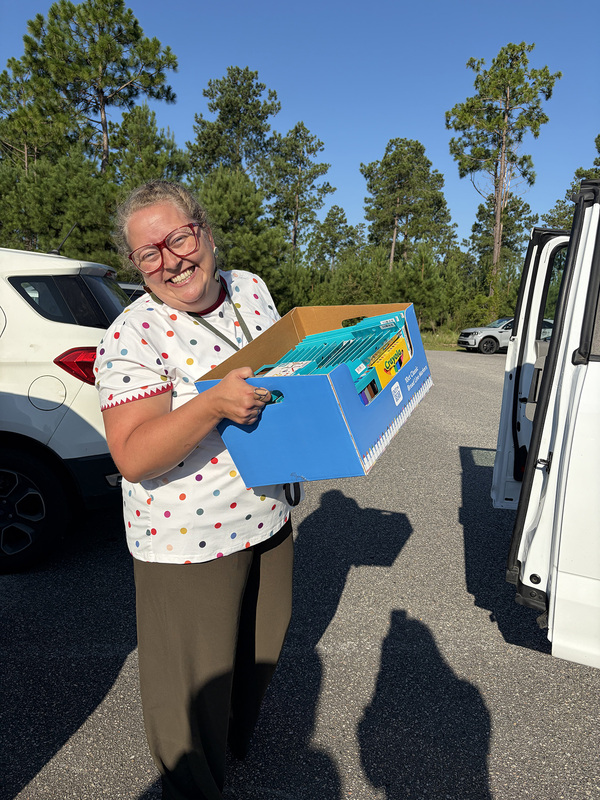 An adult carrying a box of school supplies to a vehicle.